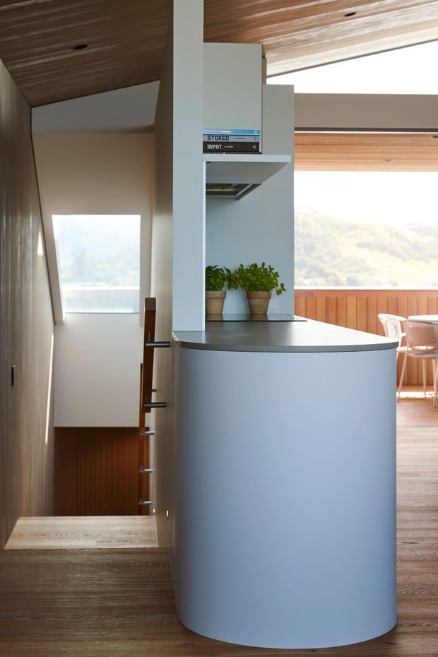 Interior view of a compact open-plan living and dining space centred on a curved pale-blue built-in island or bar with integrated shelving. The space features extensive warm timber lining on the ceiling and floors, timber wall panelling, a sculptural stair descending beside the joinery, and large glazed openings overlooking a hilly landscape. Dining furniture is visible in the background.