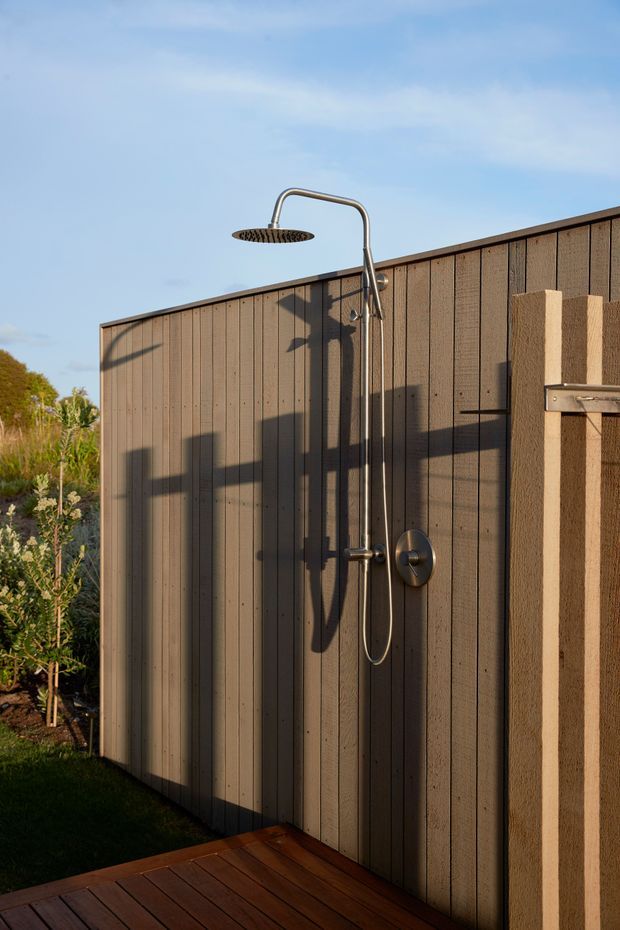 An outdoor shower mounted against a tall vertical-panel wall, with a curved brushed-metal shower arm and round showerhead. The space includes a timber deck, a textured rendered privacy wall, and landscaped planting at the edge. Warm low-angle sunlight creates strong shadows, with a clear blue sky visible above.