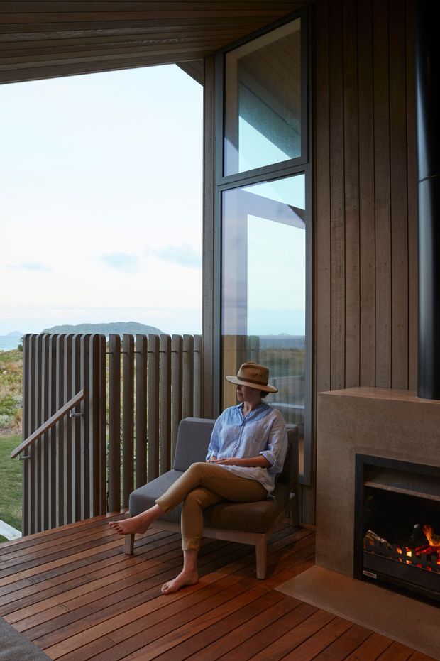 A warm, intimate interior corner beside a large glazed opening, with a person seated in an armchair next to a built-in fireplace. The space has dark timber wall and ceiling lining, timber decking-style flooring, a vertical timber privacy screen, and expansive views toward a coastal or hilly landscape. Natural light and the lit fire create a relaxed, cozy atmosphere.