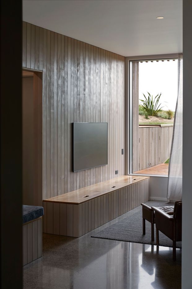 A refined contemporary living room with extensive vertical timber wall panelling and matching built-in media cabinetry. A wall-mounted television sits above a long timber console, while a large glazed opening leads to a timber deck and planted exterior area. The room includes a dark leather armchair, grey rug, polished concrete floor, recessed ceiling light, and sheer curtains. The composition emphasizes warm timber joinery, natural light, and a strong indoor-outdoor connection.