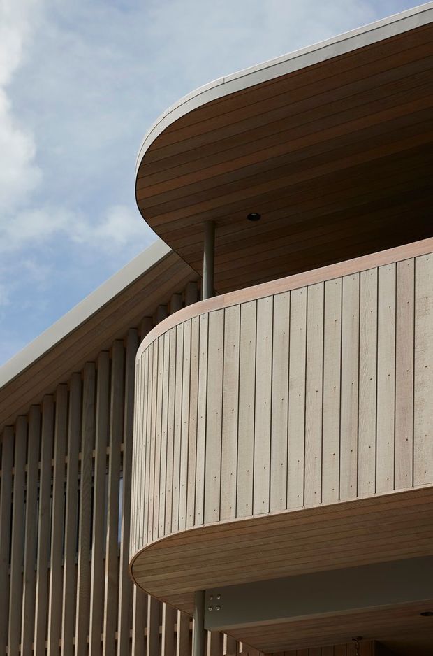 Upward-looking architectural exterior detail showing a contemporary multi-level facade with strongly rounded balcony edges, pale vertical cladding, timber-lined soffits, slender dark support columns, and a curved roofline against the sky.