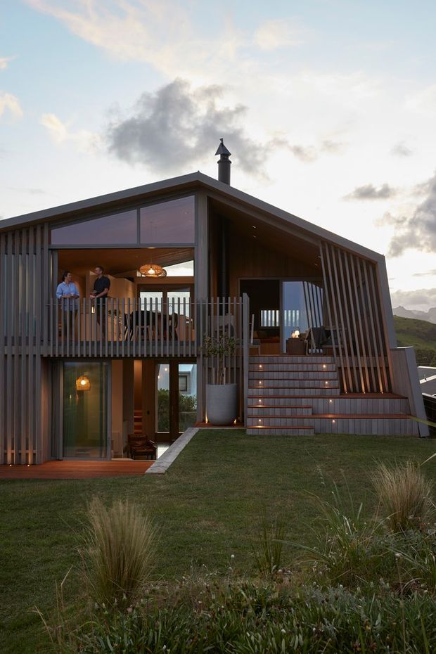 Exterior view of a contemporary two-storey house set into a landscaped sloping site. The building has a dramatic asymmetrical pitched roof, extensive glazing, dark timber cladding, vertical timber screening, an upper balcony, and broad tiled exterior stairs leading to an open living area. Warm interior lighting contrasts with the dusk sky, with mountainous terrain visible in the background.