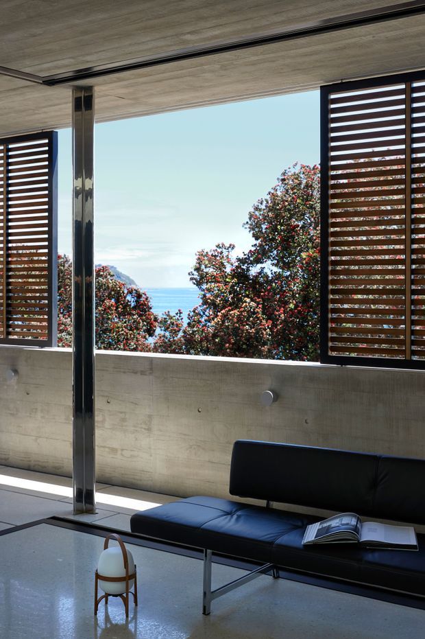 A modernist lounge or living area with a black leather sofa, low glass-topped coffee table, polished concrete floor, exposed concrete ceiling and wall, and a reflective metal structural column. Large open window bays are fitted with dark timber horizontal louvres, framing views of the ocean and coastal vegetation. The space has strong indoor-outdoor connection, abundant daylight, and a restrained material palette.
