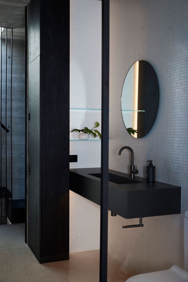 A contemporary bathroom viewed through a dark-framed glass partition. The room features small white mosaic wall tiles, a floating matte-black vanity with integrated basin, black curved tapware, an oval mirror, dark timber storage cabinetry, glass shelving, and a partially visible toilet. The space has a moody, refined palette with warm concealed lighting and a polished minimalist fit-out.