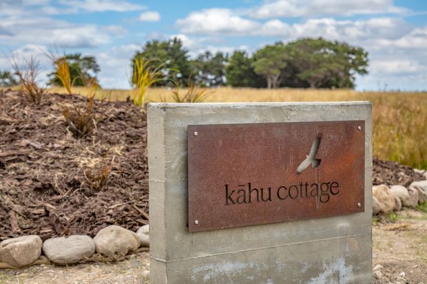 Exterior entrance sign reading “Kāhu cottage,” mounted on a weathered concrete plinth with a rusted metal plaque and bird-shaped cutout. The sign sits beside a landscaped garden bed with native-style grasses, rocks, and an open rural landscape beyond under a partly cloudy sky.