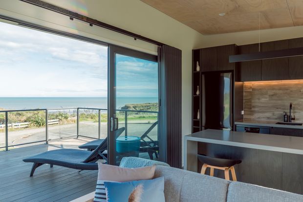 A bright open-plan coastal interior combining a compact kitchen and living area, with floor-to-ceiling sliding glass doors opening to a timber deck and ocean view. The kitchen features dark wood cabinetry, a pale stone island, a timber-toned backsplash, and warm under-cabinet lighting. A light upholstered sofa, outdoor loungers, and glass balustrade are visible, with a timber-lined ceiling adding warmth.