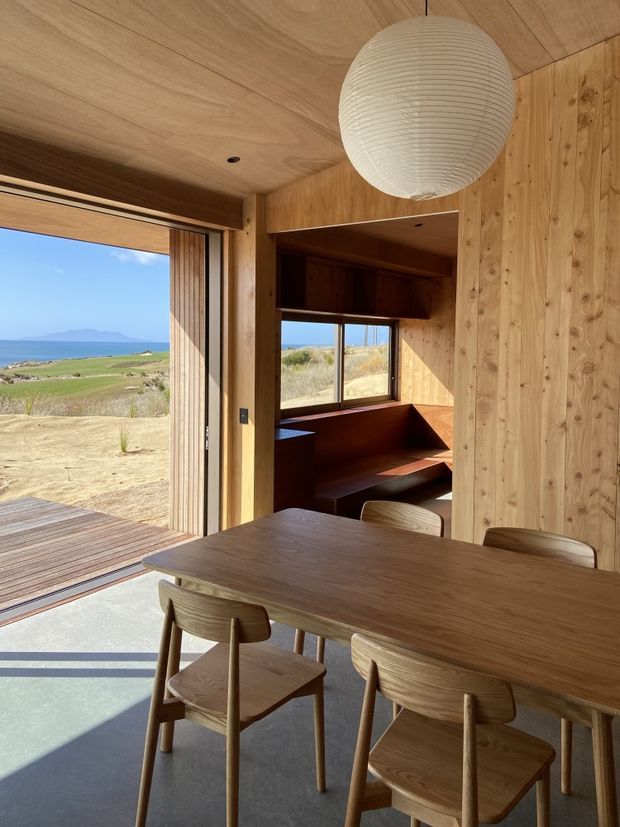 A warm timber-lined dining area with a solid wood dining table and matching wooden chairs, opening through large sliding glass doors onto a timber deck and expansive coastal landscape. Built-in timber shelving and seating occupy the adjoining space, while a large paper pendant light hangs from the timber ceiling. The room has strong indoor-outdoor connection, abundant natural light, and views toward the sea and distant hills.