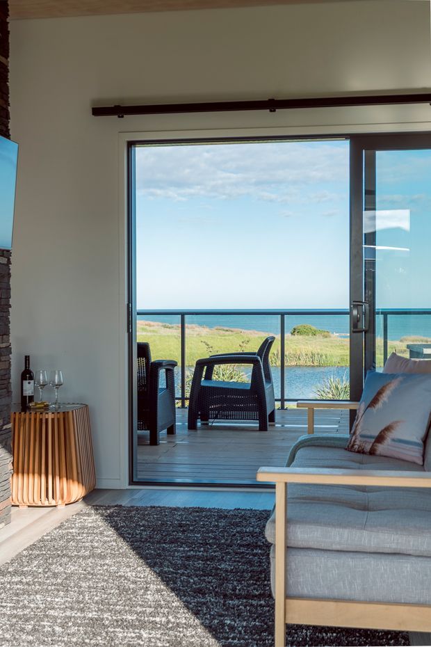 A bright coastal living room opening onto a timber deck through large sliding glass doors. The room features a light upholstered sofa with timber frame, dark textured rug, a sculptural timber slatted side table, exposed stone wall edging, and a wide ocean-facing view with outdoor seating beyond the glazing.