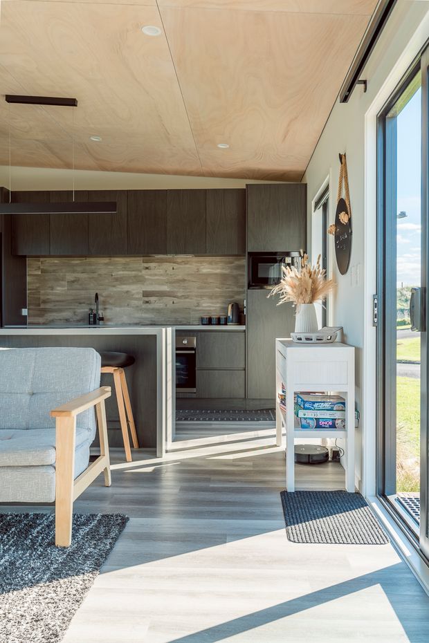 Bright compact open-plan living and kitchen space with dark wood cabinetry, a timber-lined ceiling, wood-look splashback, grey flooring, and large glazed sliding doors opening to the outdoors. A pale upholstered armchair, small white storage unit, rug, and simple pendant-style lighting create a contemporary, warm, minimalist interior.