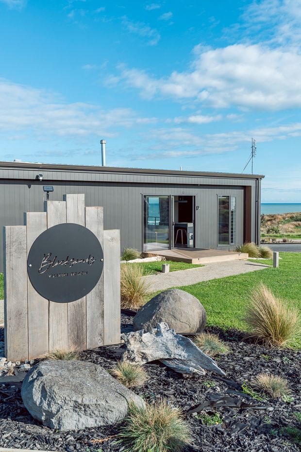 Exterior view of a low, modern coastal building with dark grey vertical cladding, a simple flat roof, glazed entrance, and a landscaped frontage. A timber monument sign stands among large rocks, black mulch, and ornamental grasses. The ocean is visible beyond the building, reinforcing the coastal setting.