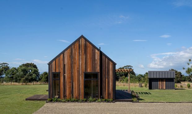 Exterior view of a compact barn-like building with a steep gabled roof and vertical dark timber cladding. Large glazed openings flank the front facade, with a small timber deck and landscaped lawn surrounding it. A separate dark-clad outbuilding is visible to the right, set in a rural-looking open landscape under a clear blue sky.