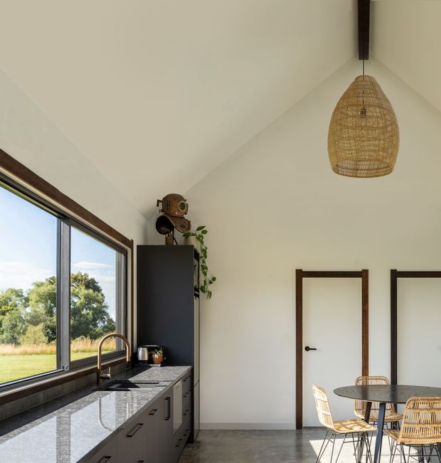 A bright, compact open-plan kitchen and dining area with a high pitched ceiling. The kitchen has dark grey cabinetry, a polished dark stone benchtop, a tiled backsplash, timber-framed glazing, and a large window overlooking greenery. A round dining table with woven chairs sits beside two white doors with dark timber trim. A woven pendant light and vintage-style decor add warmth.