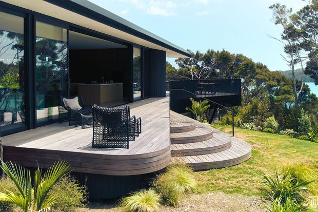 A contemporary single-storey home with dark vertical cladding and expansive black-framed glazing opens onto a raised timber deck. The deck features curved tiered steps, woven outdoor lounge chairs, and a built-in outdoor kitchen or bar area, overlooking a landscaped coastal or bush setting.