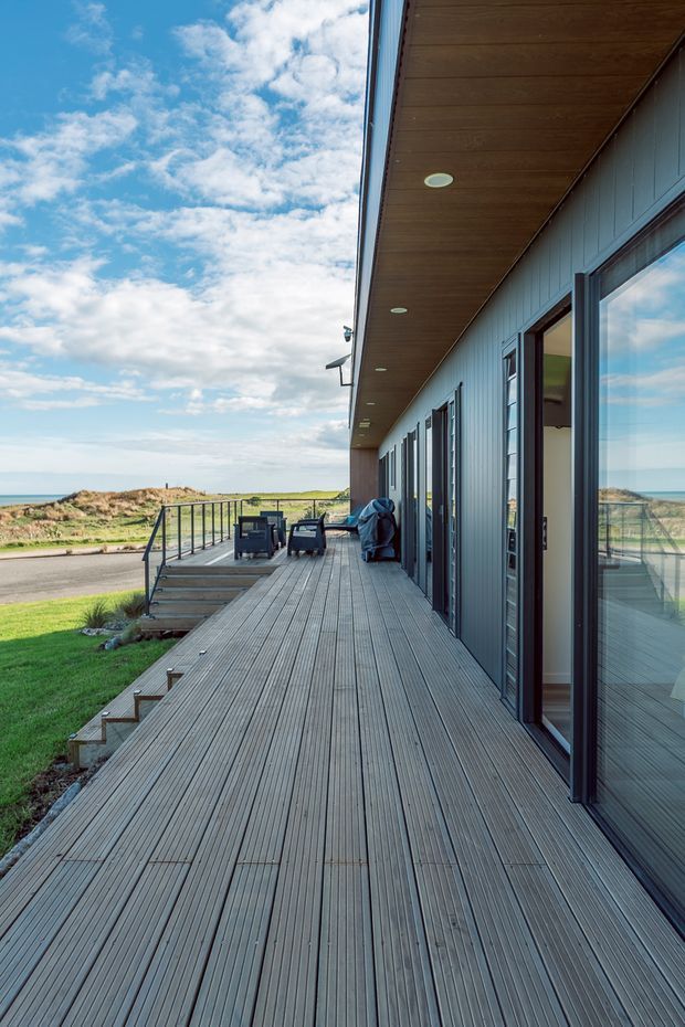 Exterior view of a contemporary coastal building from a long raised timber deck. The facade is clad in dark grey vertical panels with extensive glazed doors and windows, while the deck has timber boards, simple black metal balustrades, recessed soffit lighting, and outdoor lounge furniture. Grass, dunes, and the ocean are visible beyond the deck under a partly cloudy sky.