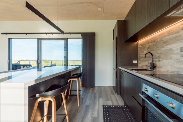 Contemporary open-plan kitchen with a large white island and integrated seating, dark timber cabinetry, dark stone-look benchtops, a textured stone-look splashback, and built-in stainless-steel appliances. A full-height glazed opening connects to a balcony or outdoor view, while timber ceiling panels and linear lighting add warmth.