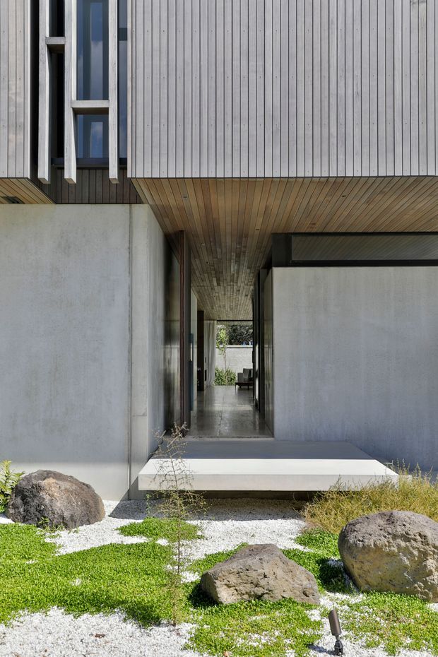 A contemporary residential entrance and covered passage framed by smooth concrete walls, vertical timber cladding, and a timber-lined soffit. The recessed entry leads through the building toward a bright courtyard, with a minimalist rock garden of gravel, grasses, groundcover, and large boulders in the foreground.
