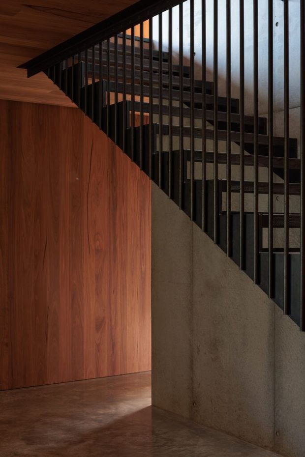 A dramatic interior stairway with a dark metal vertical-bar balustrade and exposed stair treads. Warm timber wall panelling and ceiling boards contrast with a raw grey concrete wall and polished brown concrete floor. The composition focuses on the underside and diagonal geometry of the staircase, with a moody, refined architectural atmosphere.