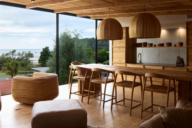 Warm contemporary open-plan kitchen and dining area with extensive floor-to-ceiling glazing overlooking a leafy coastal landscape and water. The space features a timber-lined ceiling, timber flooring, a timber kitchen with open shelving, a long island or bar counter, sculptural woven pendant lights, and timber dining stools and chairs. Soft upholstered lounge seating sits in the foreground, creating a relaxed connection between the kitchen, dining, and living zones.