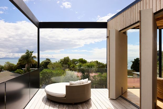 A contemporary elevated outdoor terrace with a curved upholstered daybed, timber decking, frameless glass balustrade and bold black steel framing. Vertical timber cladding and a large open pivot door connect the terrace to the interior, with expansive coastal and water views beyond.