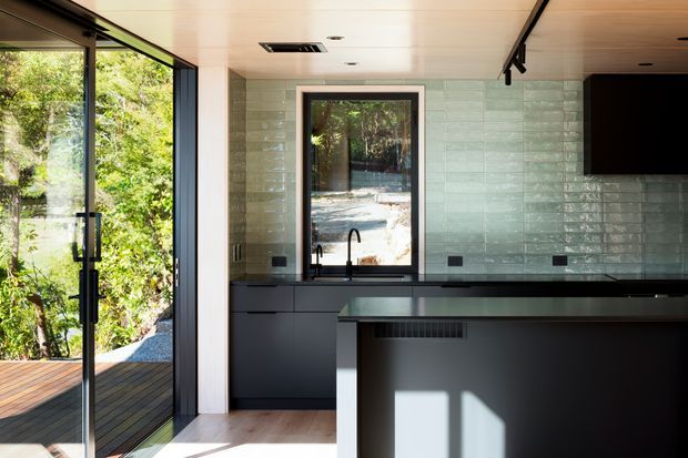A contemporary kitchen with dark matte cabinetry and a large island, set against a pale green glossy rectangular-tile splashback. Full-height black-framed glazing and a glazed door open toward a timber deck and leafy garden. The kitchen has a light timber ceiling, pale timber flooring, integrated appliances, and a wall-mounted black screen.