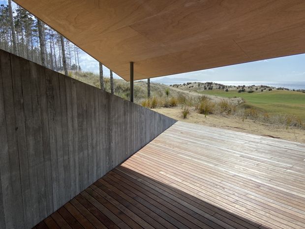 A minimalist covered outdoor deck or terrace overlooking a grassy coastal landscape. The space features warm timber decking and a broad flat timber soffit supported by a slender post, with a dark vertical timber privacy wall forming one side. The open edge frames views toward dunes, vegetation, and the distant sea.