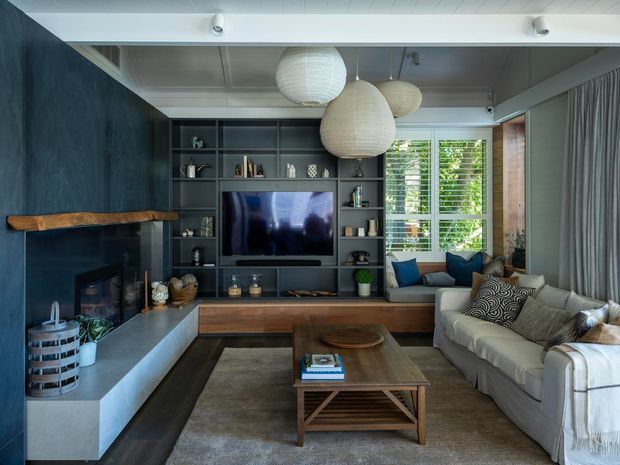A compact, sophisticated living room with a dark blue textured feature wall, built-in charcoal shelving and television cabinetry, a linear fireplace with a timber mantel, and a low timber bench beneath the shelving. A pale upholstered sofa with patterned cushions faces a timber coffee table, while large woven pendant lights hang from a white paneled ceiling. Shuttered windows bring in filtered daylight, and full-height curtains frame the room.