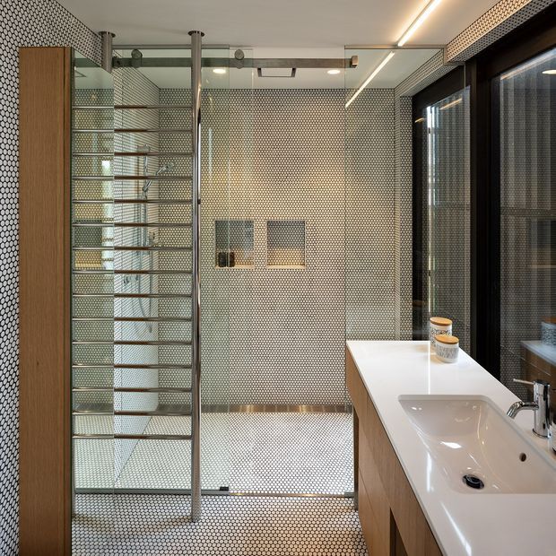 Contemporary bathroom with a walk-in shower enclosed by clear glass and framed by slim metal posts, a wall-hung shower fixture, and extensive small-format patterned tile covering the walls and floor. A long white integrated vanity with a built-in basin and timber-look cabinetry runs along the right side, beside dark-framed glazing. The space is bright, refined, and highly coordinated in neutral tones.