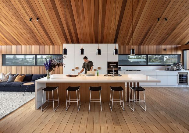 Open-plan contemporary kitchen with a large white island and five black bar stools, integrated white cabinetry, timber wall and ceiling panelling, and warm timber flooring. A lounge area is visible to the left, while broad horizontal windows bring in natural light. Black pendant lights and recessed ceiling lights provide contrast against the warm timber interior.