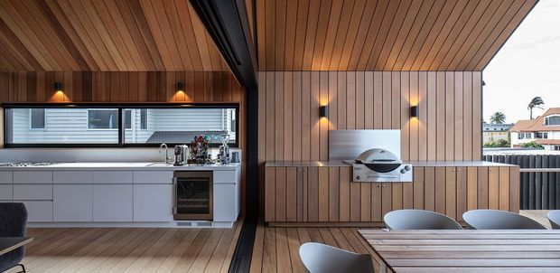 A refined outdoor entertaining area with a built-in barbecue and dining table, connected to a compact linear kitchen. Warm timber slat cladding continues across the walls and angled ceiling, contrasted with white cabinetry, a pale benchtop, dark metal framing, and integrated wall lighting. The space is sheltered yet open to the surrounding suburban setting.