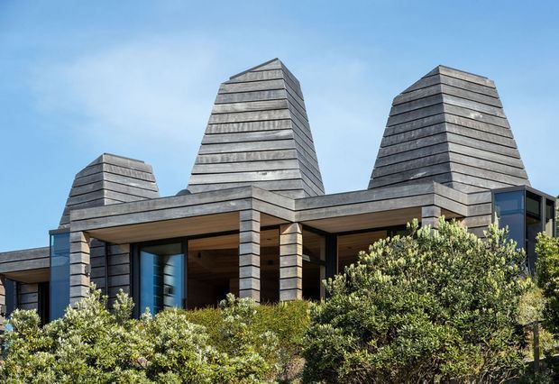 Exterior view of a contemporary, low-rise residence with three prominent tapered chimney-like tower forms and extensive horizontal grey timber cladding. Deep roof overhangs and recessed glazed openings create sheltered outdoor-facing spaces, while dense native-looking shrubs partially screen the lower facade. The composition is set against a clear blue sky.