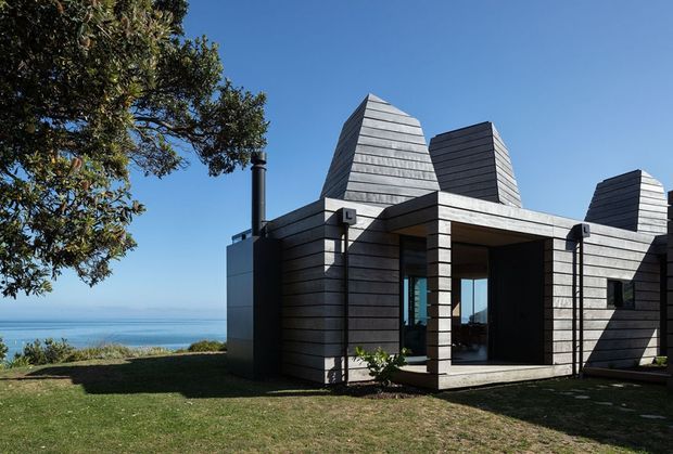 A modern coastal house viewed from the lawn, clad in horizontal grey weatherboards with several distinctive steep, faceted roof forms. Deep recessed openings and large glazed doors create a sheltered outdoor-facing entry or living area. The property sits beside a broad blue coastal or ocean outlook, with mature landscaping and strong clear daylight.