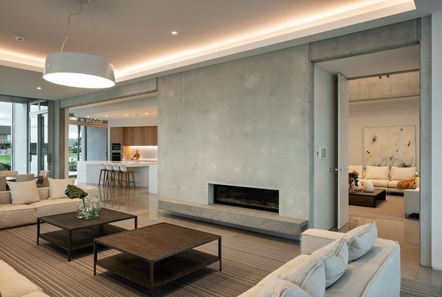Open-plan contemporary living room with a concrete feature wall and built-in linear fireplace, large neutral sofas, dark wood-and-metal coffee tables, and a striped area rug. A modern kitchen with island seating is visible in the background, while an adjacent bedroom or sitting area is visible through the doorway. Recessed and cove lighting create a refined, warm atmosphere.