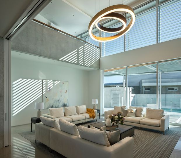 A bright, spacious contemporary living room with a double-height volume, large glazed openings, and a sculptural circular pendant light. Multiple cream sectional sofas surround a low coffee table, while strong striped sunlight from the horizontal louvres creates dramatic patterns across the walls and floor. Concrete surfaces, slim metal-framed glazing, and an upper-level balustrade contribute to the modern architectural character. A pool and exterior structures are visible beyond the glazing.