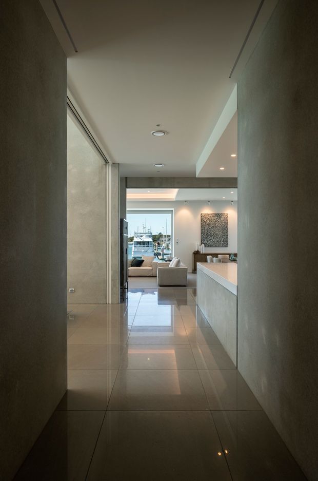 A long contemporary interior corridor leading into an open-plan living and kitchen area. The space features large-format glossy beige stone or porcelain floor tiles, smooth textured grey walls, concealed linear air-conditioning vents, recessed ceiling lights, and a pale streamlined kitchen island. A marina and boats are visible through the rear glazing, while a neutral sofa and artwork occupy the living area.