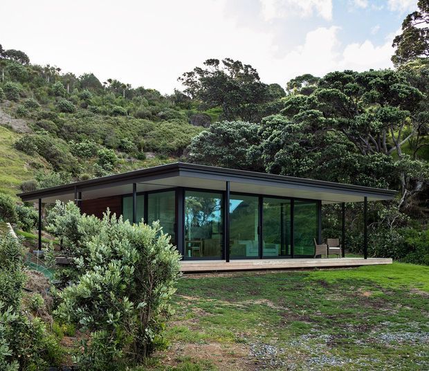 A low, single-storey modern pavilion set within dense native vegetation on a sloping site. The building has a broad flat roof with deep overhangs, dark slender framing, extensive floor-to-ceiling glazing, and a timber-toned deck extending along the facade. Interior furnishings are faintly visible through the glass, while landscaped planting and a gravel-and-grass foreground surround the structure.