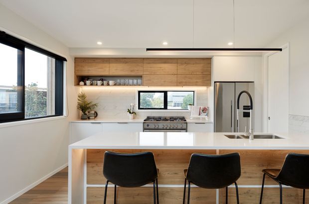 Bright contemporary kitchen with a large white island and integrated seating, white cabinetry, stainless-steel appliances, a timber feature wall cabinet, and black-framed windows and pendant lighting. The space has a clean, functional layout with warm timber accents and abundant natural light.