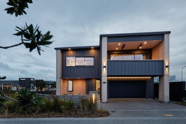 Contemporary two-storey residential facade photographed at dusk, featuring dark vertical cladding, warm timber-lined recessed areas, a projecting upper balcony, integrated lighting, and a large enclosed garage. Landscaping and neighboring buildings are visible in the foreground and background.