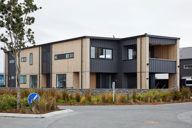 Contemporary multi-storey residential building with a strong geometric composition, combining vertical timber cladding with dark charcoal cladding. The facade includes recessed balconies, large rectangular windows, covered entry areas, and a flat roof. Low-maintenance landscaped planting and a paved street frontage are visible in the foreground.