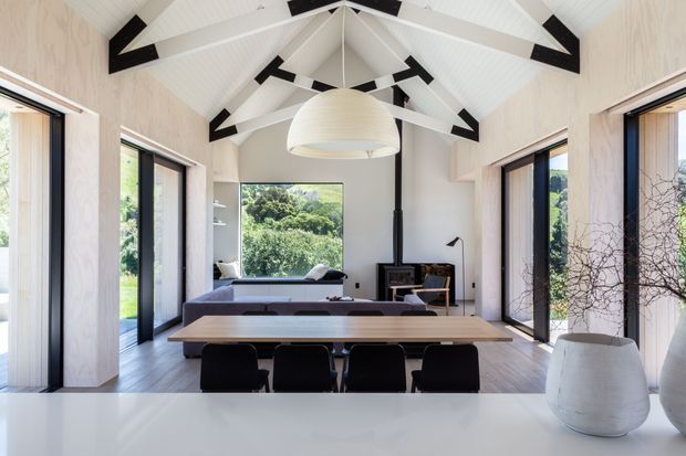 Bright contemporary open-plan living and dining space with a dramatic white vaulted ceiling, exposed black-and-white structural beams, extensive full-height glazing, a long timber dining table, and a relaxed seating area overlooking greenery. Pale timber wall panels and flooring create a warm, natural feel, while a large sculptural pendant anchors the room.