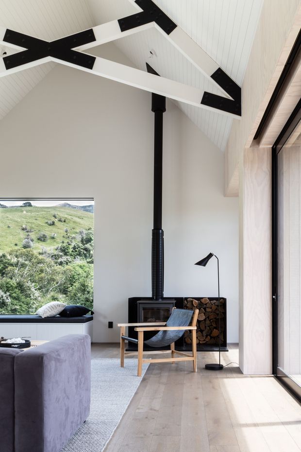 Bright contemporary living room with a dramatic high pitched white timber-lined ceiling, exposed black structural beams and a central black flue. The room includes a freestanding wood-burning stove with stacked firewood, a timber-framed lounge chair, floor lamp, sofa, pale rug and light timber flooring. Large windows provide views toward a green rural landscape, while full-height glazing along the right side brings in abundant natural light.