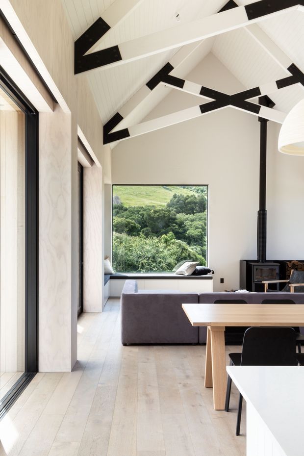 Bright contemporary open-plan interior combining a living room, dining area and kitchen. The space features pale timber flooring and wall panelling, white-painted surfaces, expansive black-framed glazing, a sculptural pitched ceiling with exposed white rafters and black connection plates, a grey sofa, timber dining table and a freestanding black wood-burning stove. Lush greenery is visible through the large window.