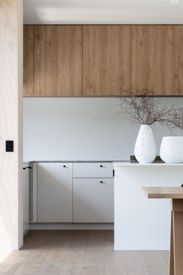 Bright, refined kitchen viewed through a wide opening, featuring full-height warm timber overhead cabinetry, white vertical-panel base cabinetry, a pale rectangular tile splashback, and a light timber island or peninsula. Neutral stone-look flooring and sculptural white vases with branches add a calm contemporary character.