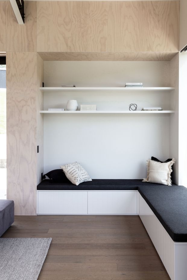 A bright, minimalist built-in window-seat nook with an L-shaped dark upholstered bench, white vertical-groove cabinetry, open shelving, and pale plywood wall and overhead joinery. The space includes a few neutral cushions, books, and simple decorative objects, with timber flooring and abundant natural light.