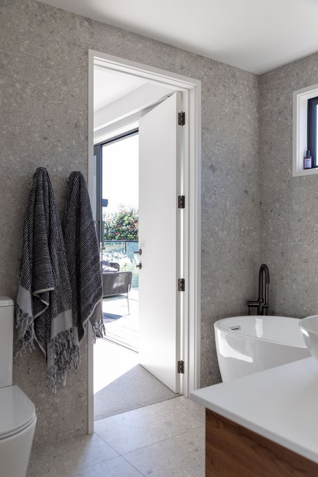 Bright contemporary bathroom featuring grey stone-look wall and floor finishes, a freestanding white bathtub, black wall-mounted tapware, white vanity benchtop with timber cabinetry, and a white toilet. An open doorway leads to an outdoor terrace or adjacent exterior area.