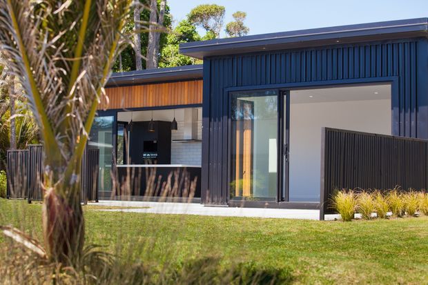 Exterior view of a contemporary single-storey home with dark vertical cladding, broad glazed sliding doors, and a partially visible outdoor kitchen or entertaining area. Warm timber cladding accents the recessed section, while landscaped lawn and tropical planting frame the facade.