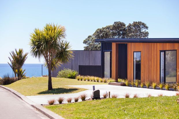A contemporary single-storey coastal house viewed from the street, with a flat roof, dark vertical cladding, warm timber-look facade panels, large black-framed windows, and a landscaped front lawn. The ocean is visible beyond the property, with palm-like plants and low ornamental planting around the house.