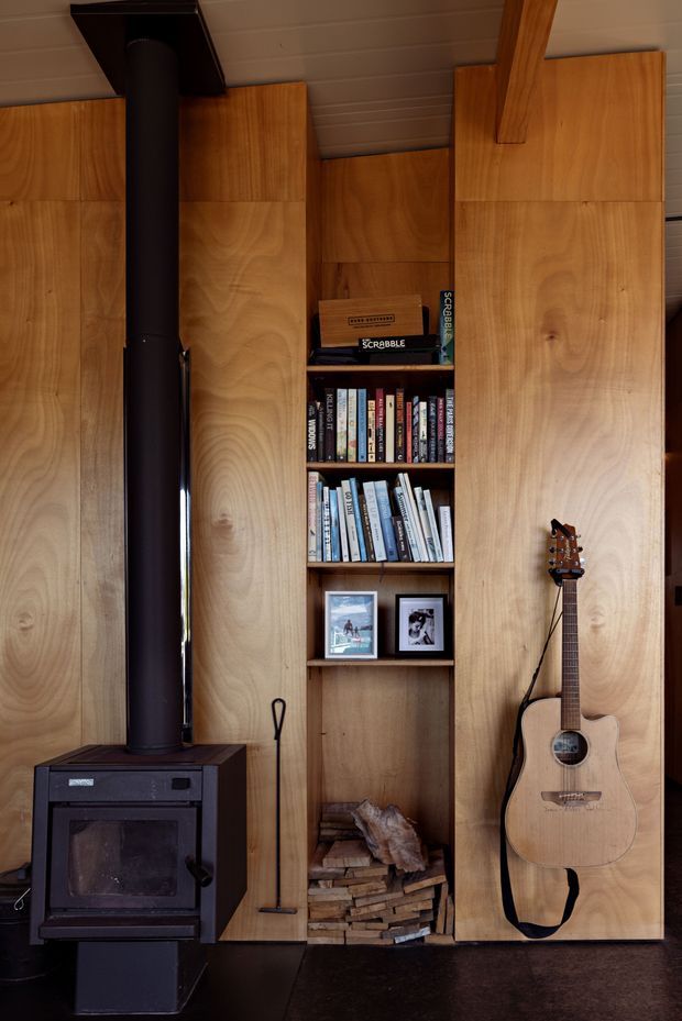 A compact, warm timber-lined interior featuring built-in shelving filled with books and board games, a black freestanding wood-burning stove with flue, stacked firewood storage, framed photos, and an acoustic guitar. The vertical timber wall panels and exposed ceiling create a cozy cabin-like character.