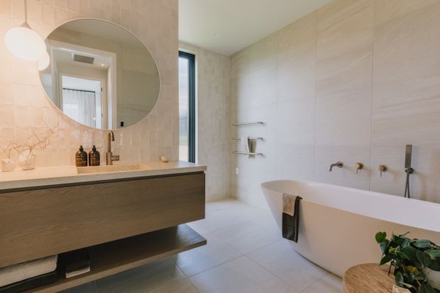 A bright, refined contemporary bathroom with a freestanding white bathtub, wall-mounted brushed-metal bath fittings, a floating timber vanity with an integrated stone benchtop, and a large round mirror. Pale stone-look wall and floor tiles create a warm, neutral palette, while a narrow vertical window and soft pendant lighting add natural light.