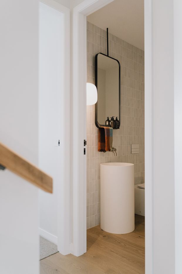 A compact contemporary bathroom viewed through an open doorway, featuring full-height beige grid-pattern ceramic wall tiles, warm timber flooring, a freestanding cylindrical white basin, and a rounded rectangular black-framed mirror with integrated shelf and pendant lighting. White door and wall trim frame the room, while a partial hallway and timber handrail are visible in the foreground.