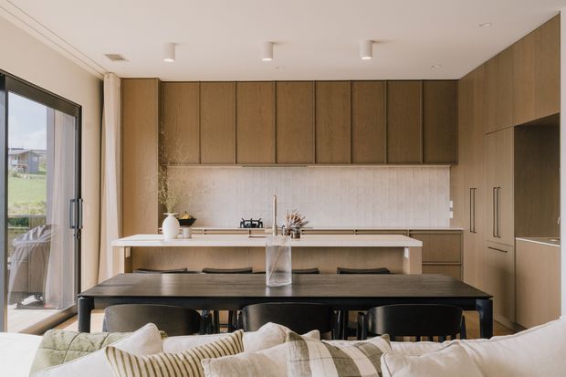 A bright, contemporary open-plan kitchen and dining area viewed from the living room. Full-height warm timber cabinetry lines the rear wall and continues into a tall bank on the right, with a pale textured tiled splashback, light benchtops, and a central island or peninsula. A dark timber dining table with black chairs occupies the foreground, while a large glazed sliding door brings in natural light and connects to the exterior.